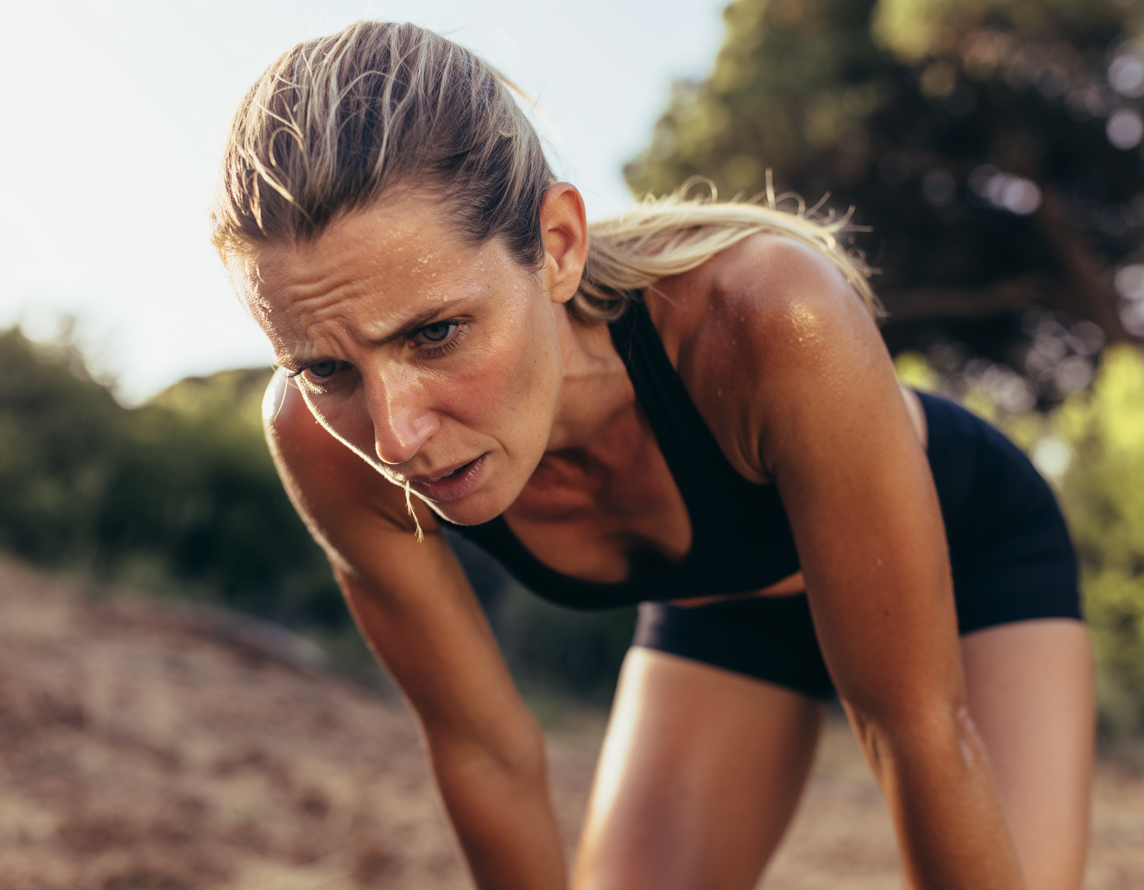 Verschwitzte Sportlerin im Freien, nach dem Training nach vorn gebeugt und erschöpft, stützt sich mit den Händen ab.