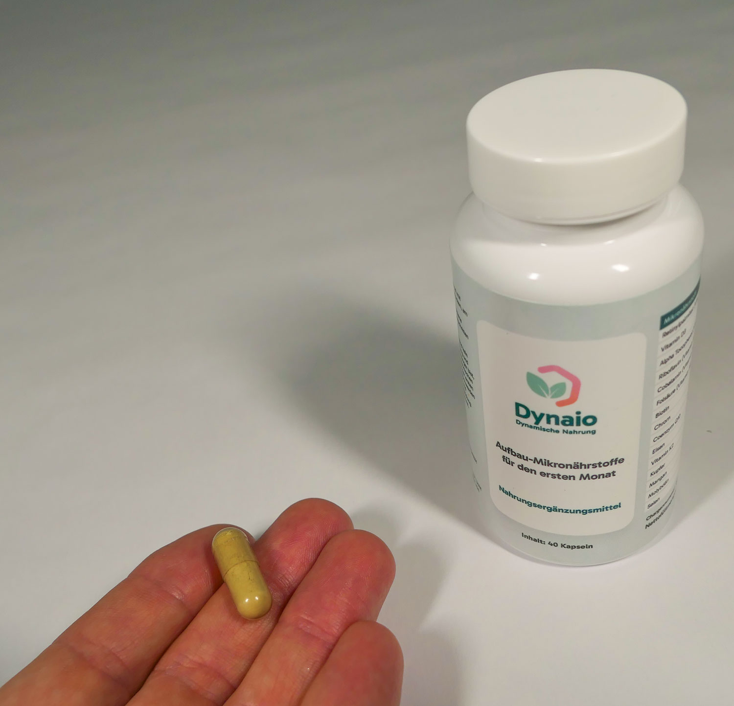 Eine Dynaio Kapsel in der Hand, Dynaio Dose rechts daneben